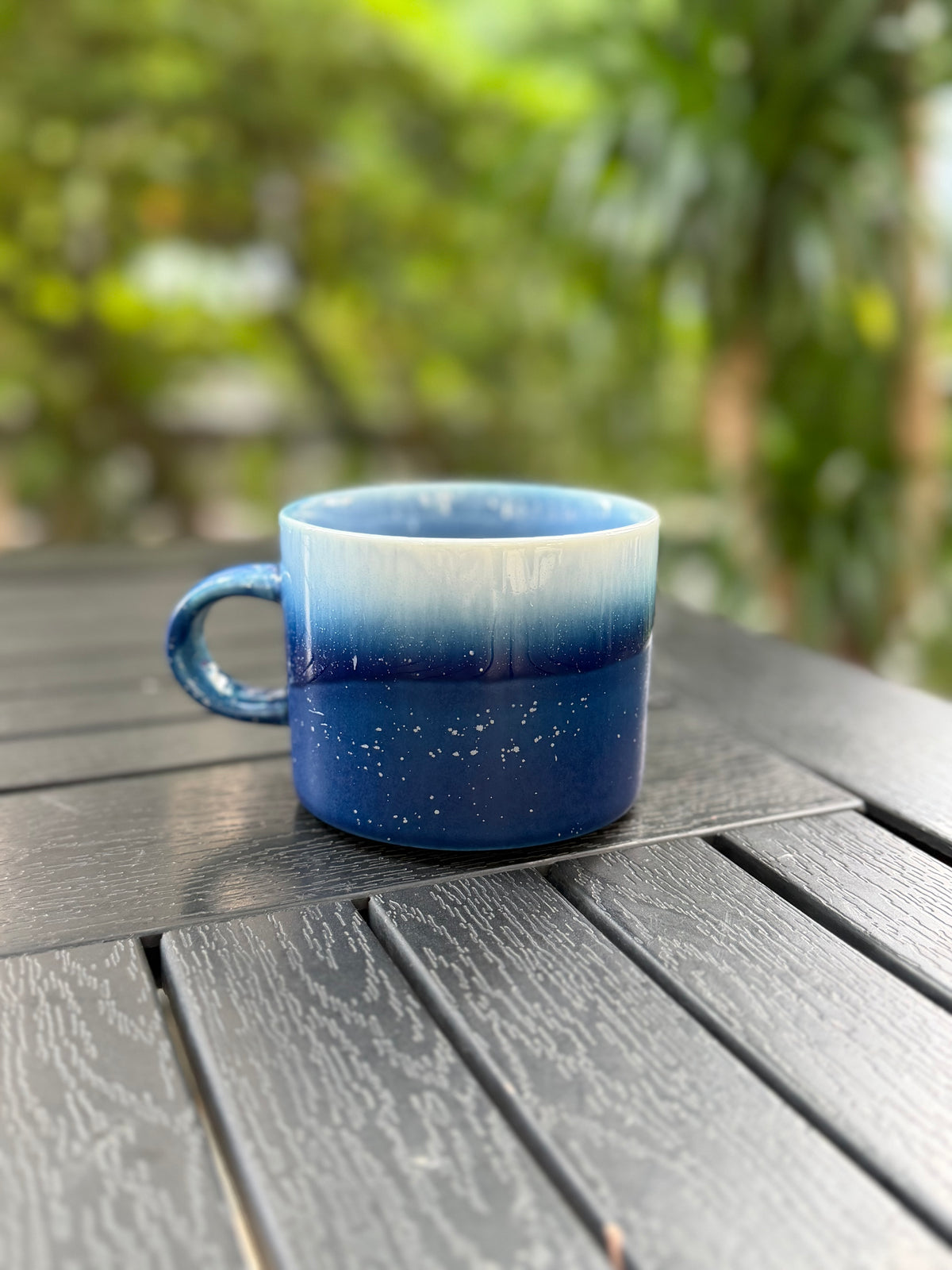 Back view of the blue kiln-glazed mug on an outdoor table with softly blurred greenery