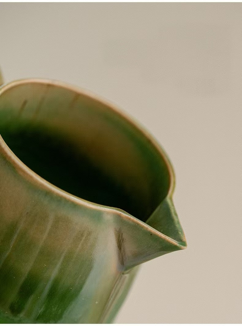 Close-up of a ceramic pitcher with a glossy green glaze and a light beige rim, shown against a neutral light gray background.