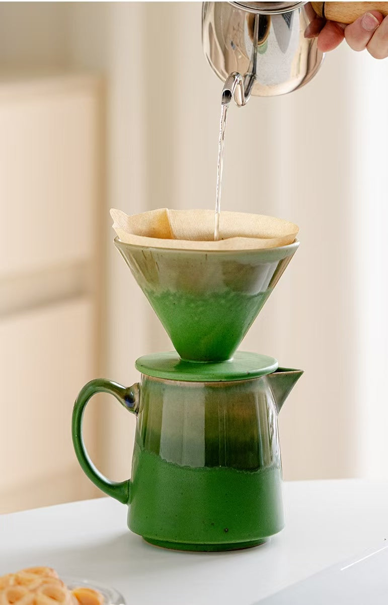 A green ceramic pour-over coffee set with a matching carafe and paper filter, shown with hot water being poured from a metal kettle. A hand is visible holding the kettle, and the scene is set on a white table with a softly blurred indoor background.