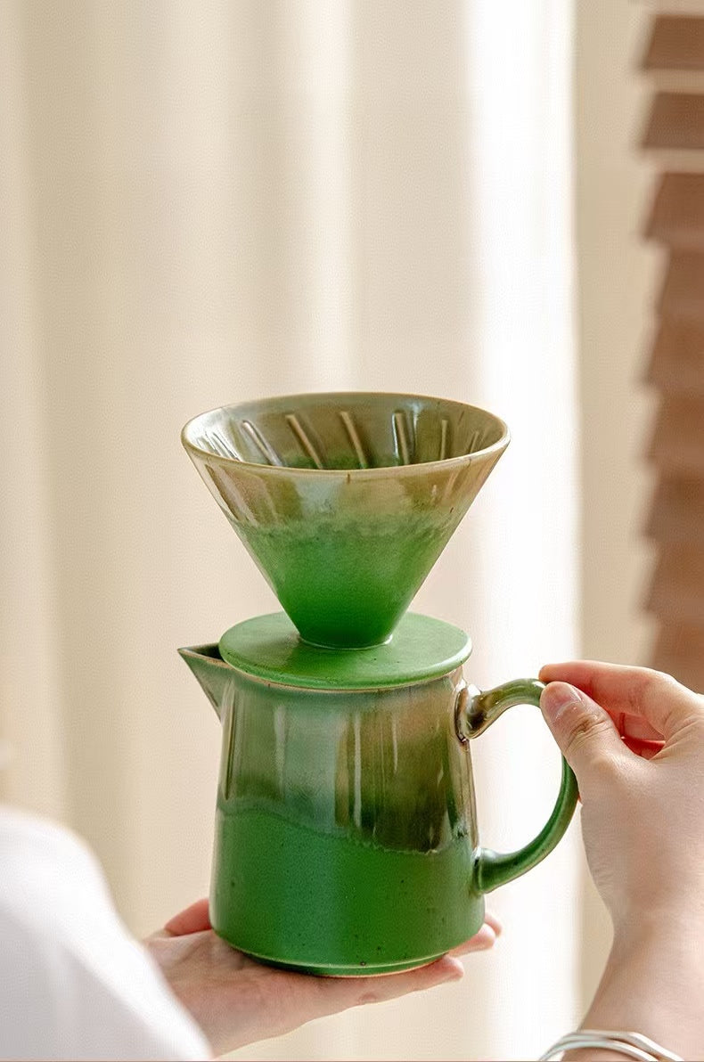 A green ceramic pour-over coffee set with a matching carafe, held by hands against a softly blurred indoor background.