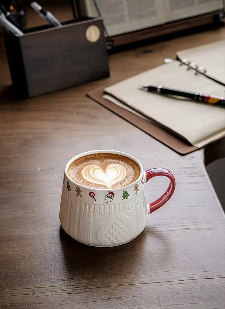 White Winter Knit holiday ceramic mug with red handle filled with latte on a wooden desk