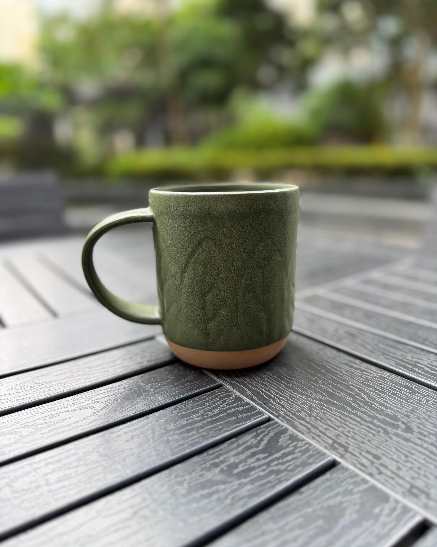 Side view of the Woodland Sprig stoneware mug on a dark slatted outdoor table, handle facing left, showing the subtle leaf emboss texture and matte forest green finish with natural stoneware base