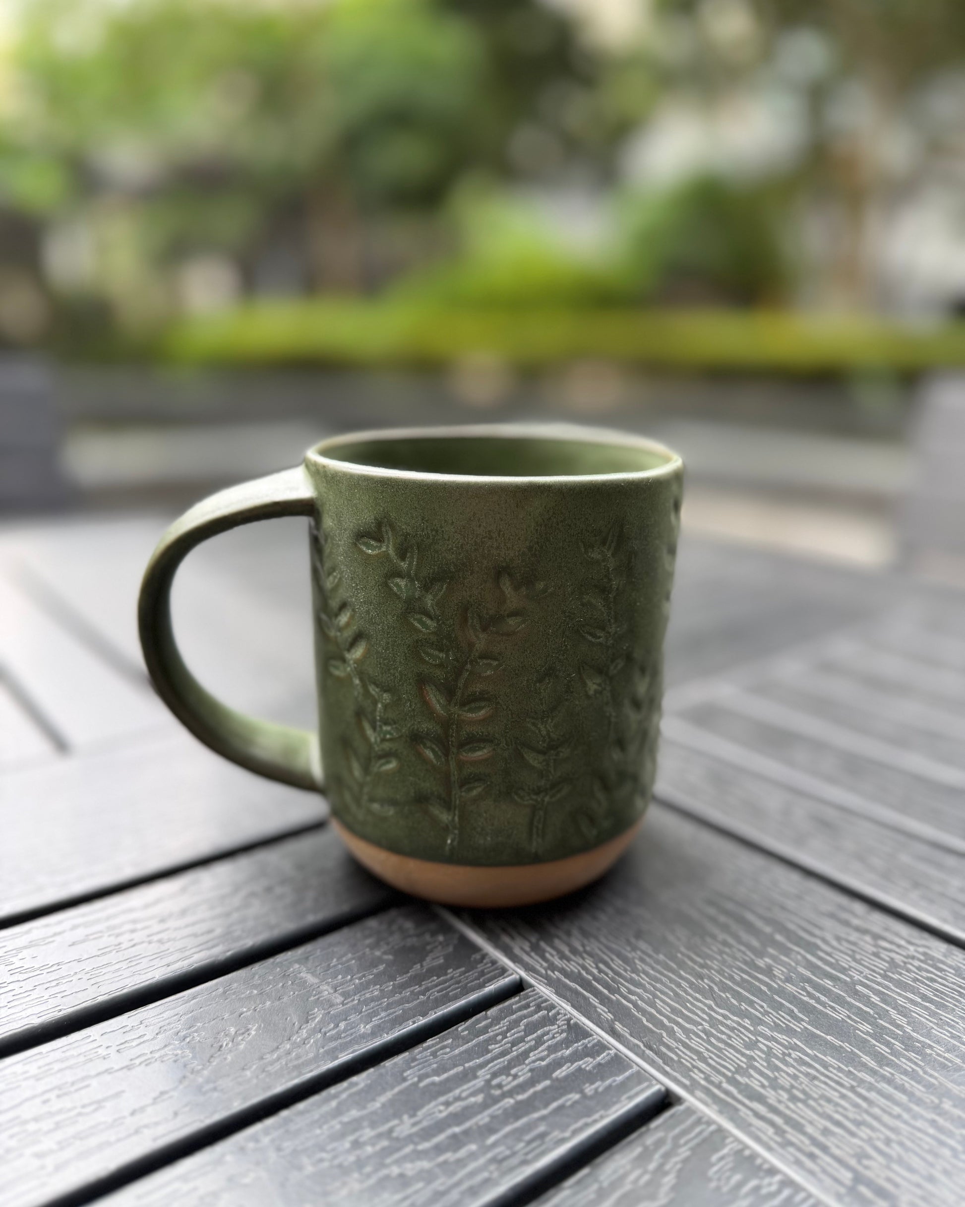 Front view of the Woodland Sprig stoneware mug on a dark slatted outdoor table, handle facing left, highlighting the all-over embossed sprig pattern and deep forest green glaze