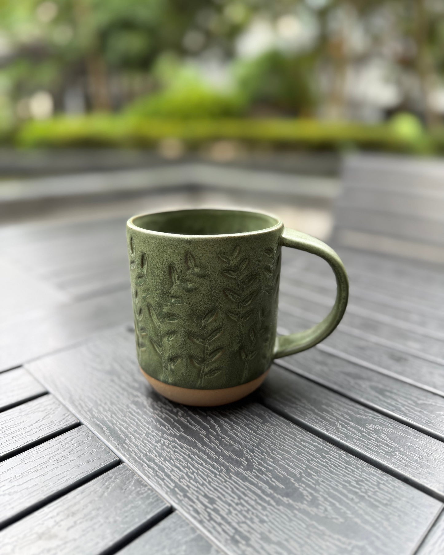 Woodland Sprig stoneware mug on a dark slatted outdoor table, handle facing right, showcasing the embossed botanical leaf relief and forest green matte glaze with exposed stoneware base