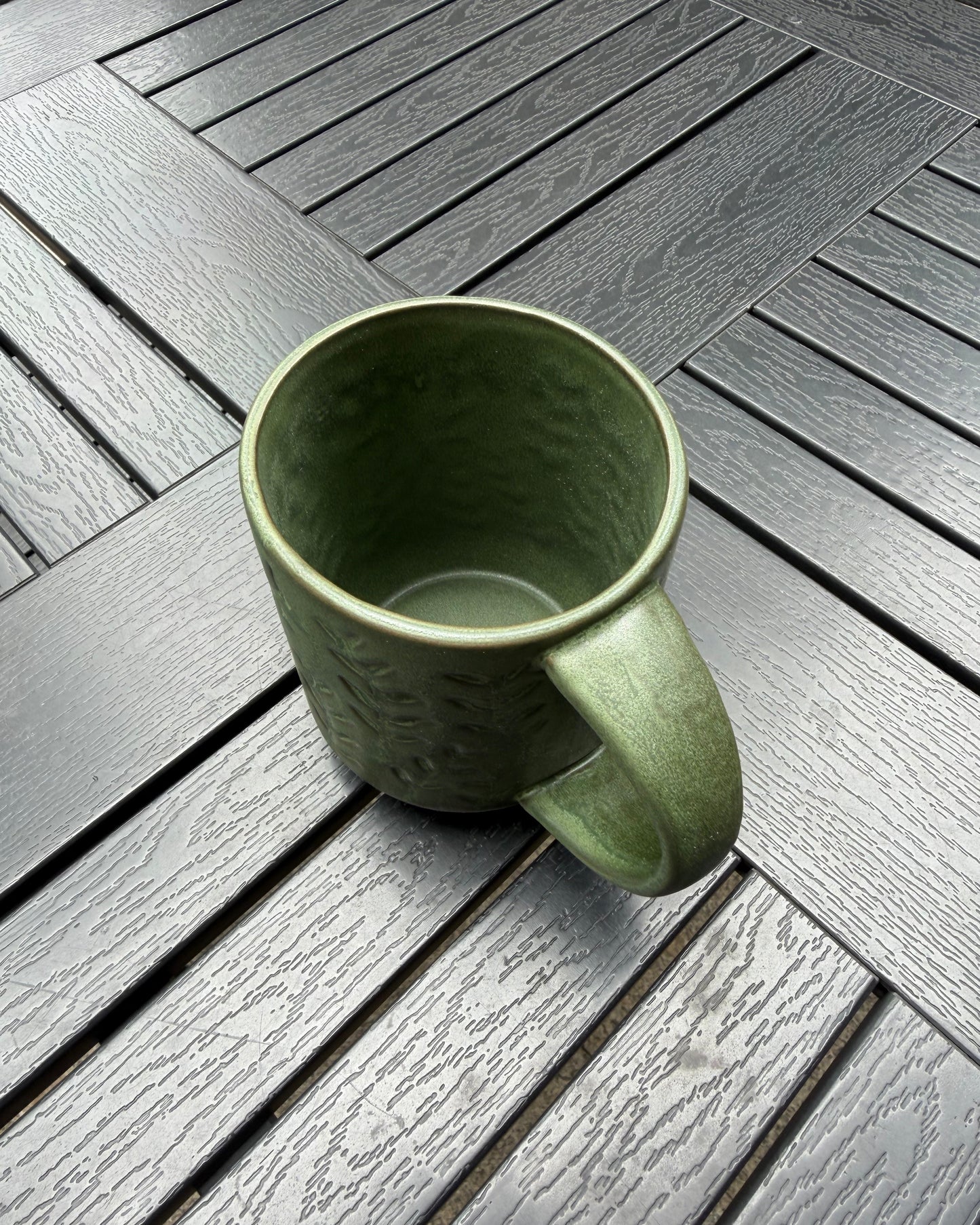 Top-down view of the Woodland Sprig stoneware mug on a dark slatted outdoor table, showing the open rim, interior glaze, and embossed botanical pattern wrapping the exterior