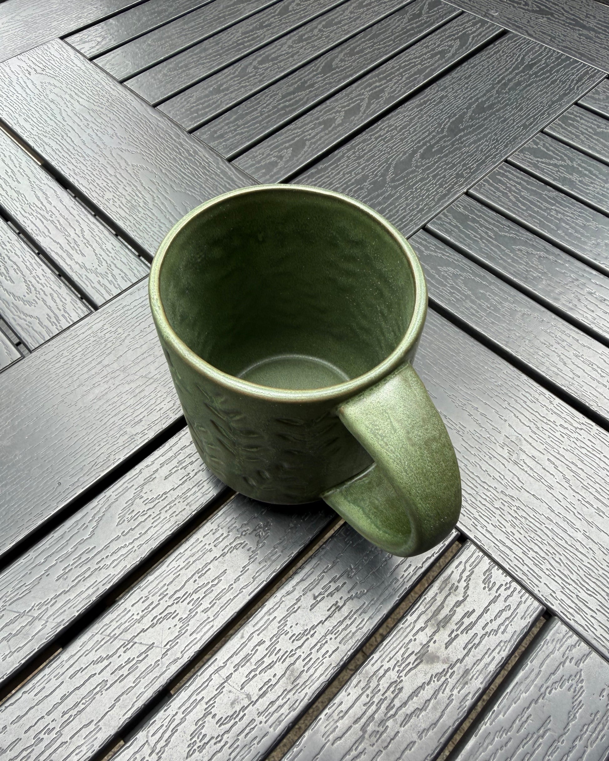 Top-down view of the Woodland Sprig stoneware mug on a dark slatted outdoor table, showing the open rim, interior glaze, and embossed botanical pattern wrapping the exterior